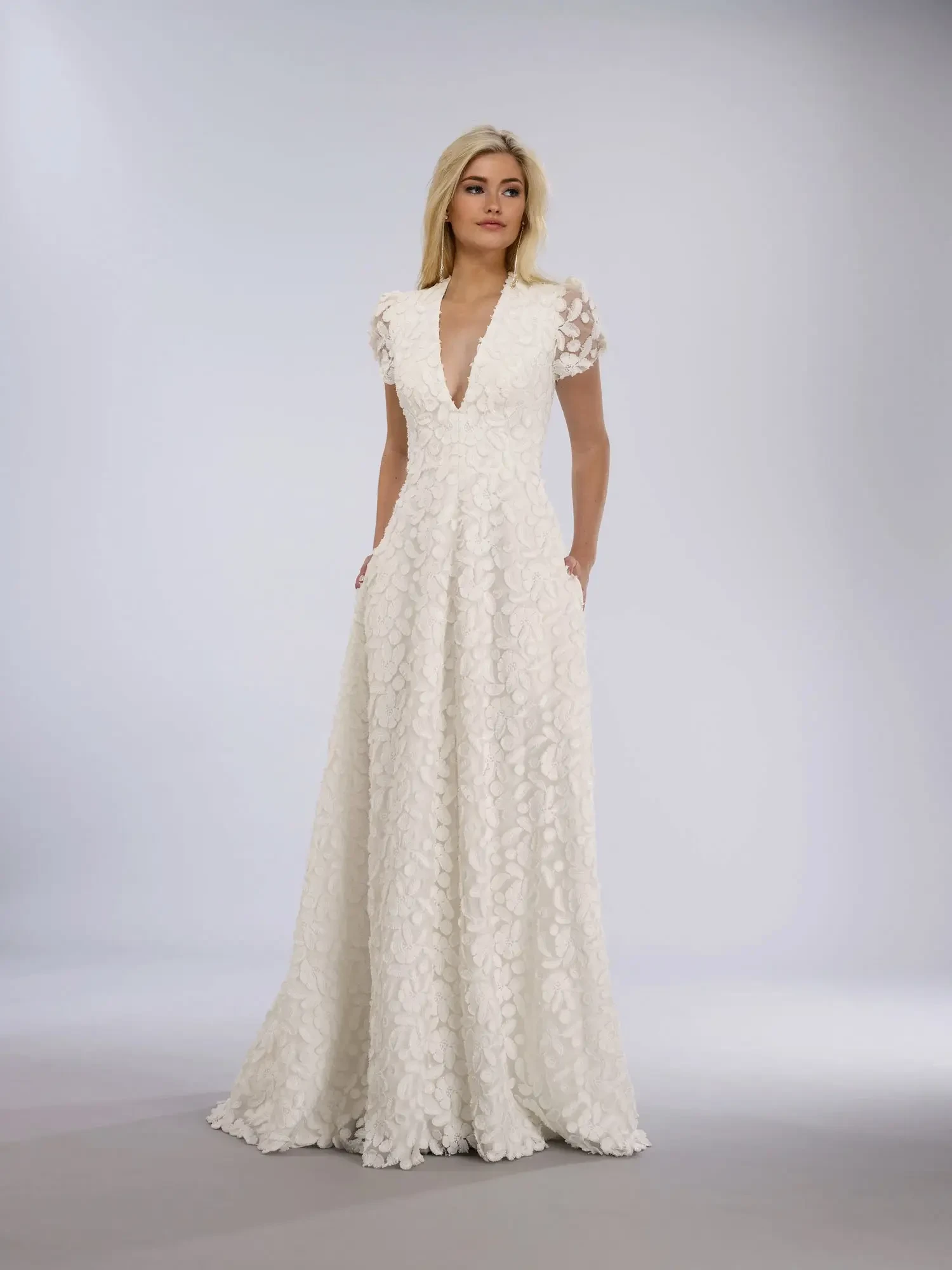 A woman poses in a white, floral-textured gown with short sleeves and a deep V-neckline. She stands confidently, conveying elegance and grace.