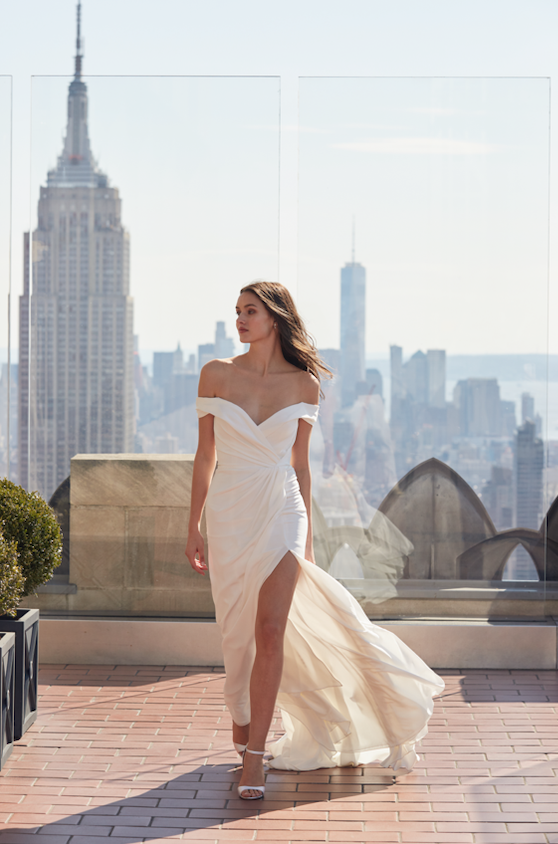 Model is wearing an off-the-shoulder fitted wedding gown with a plunging v-neckline and a thigh-high slit.
