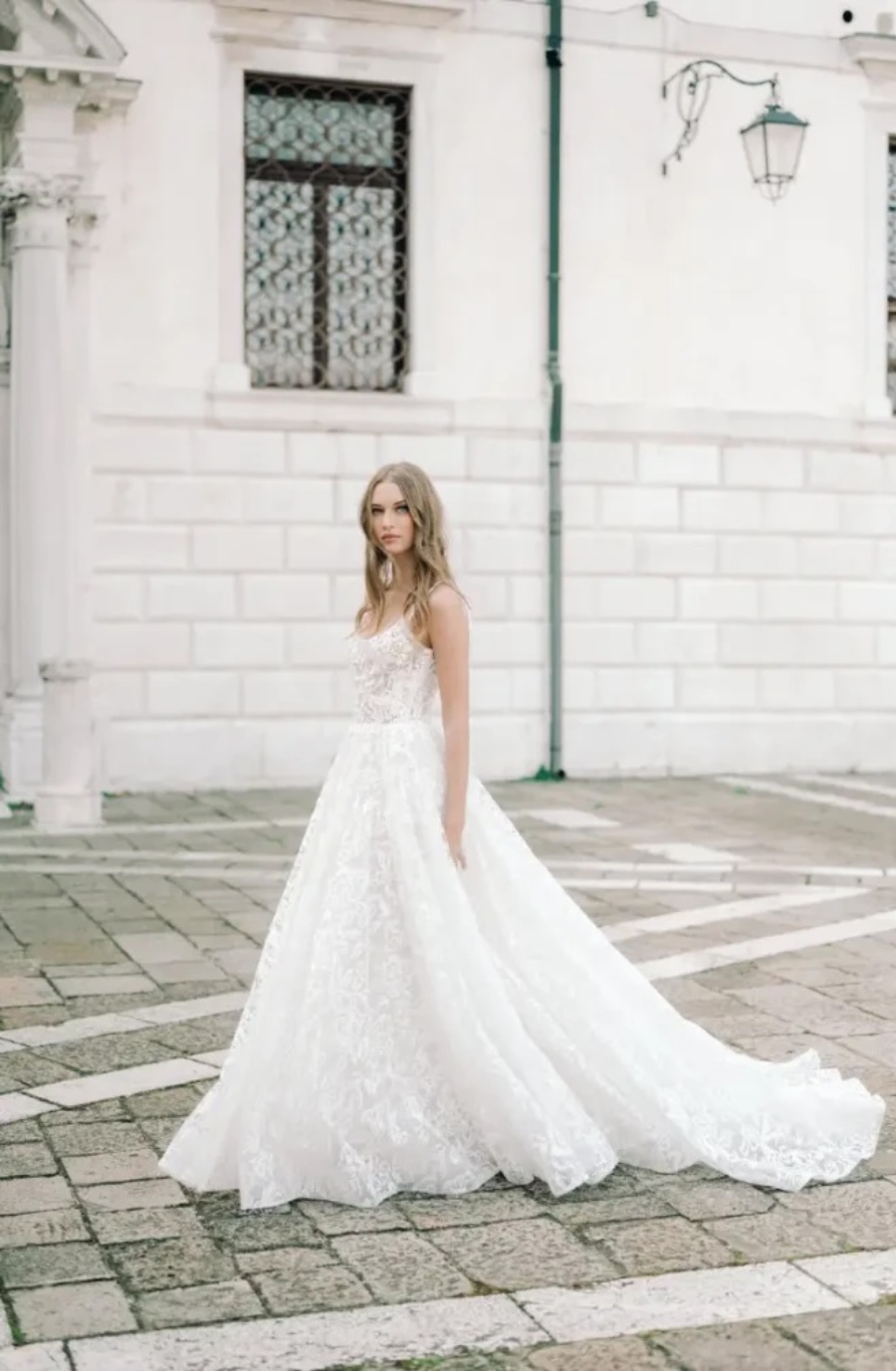 A woman in a flowing white lace wedding gown stands on a cobblestone street. The background features a historic building with ornate windows, creating a serene and elegant atmosphere.