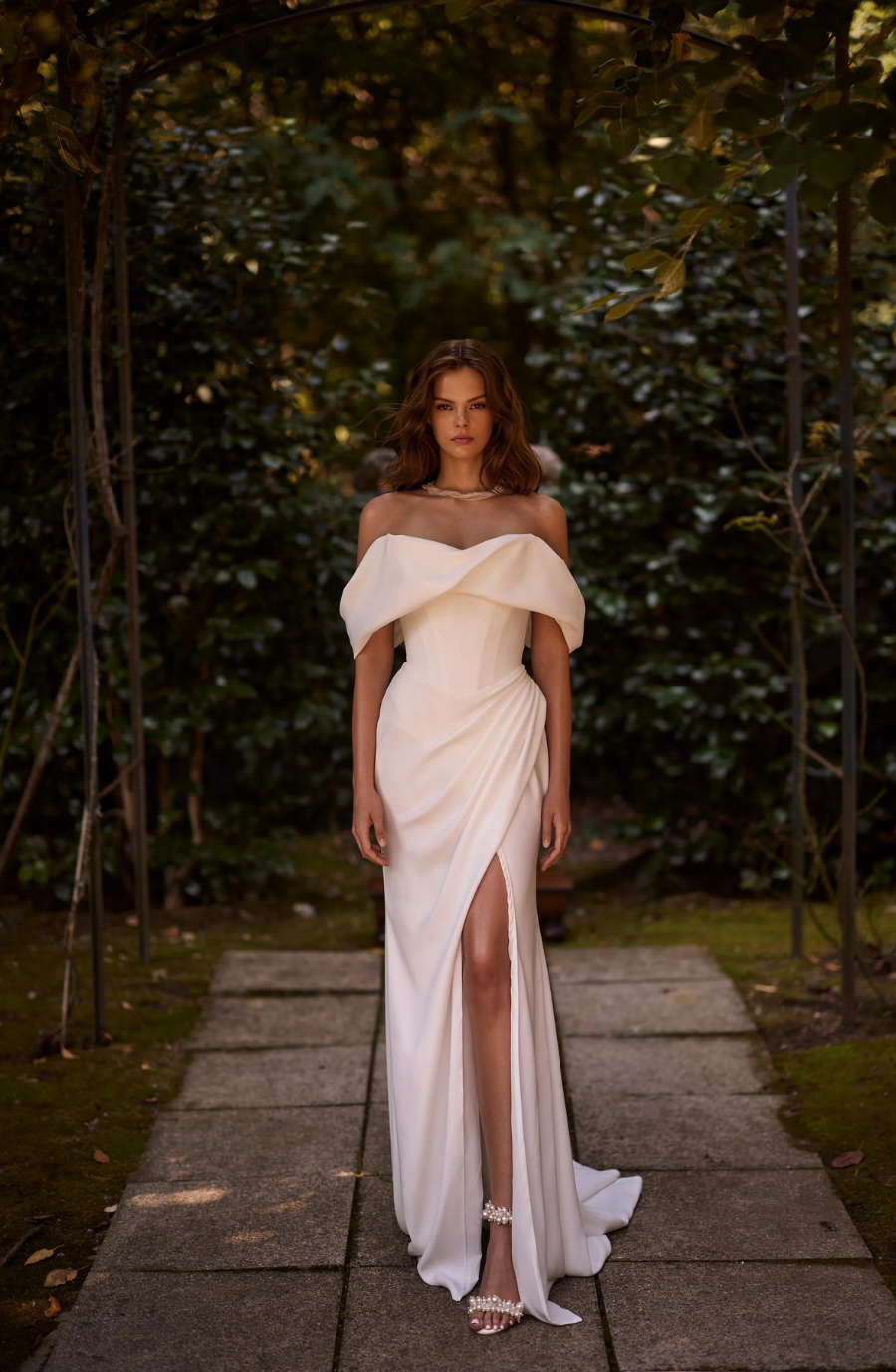 Woman in elegant off-shoulder white gown with thigh-high slit stands on garden path. Lush greenery surrounds, creating a serene and sophisticated tone.