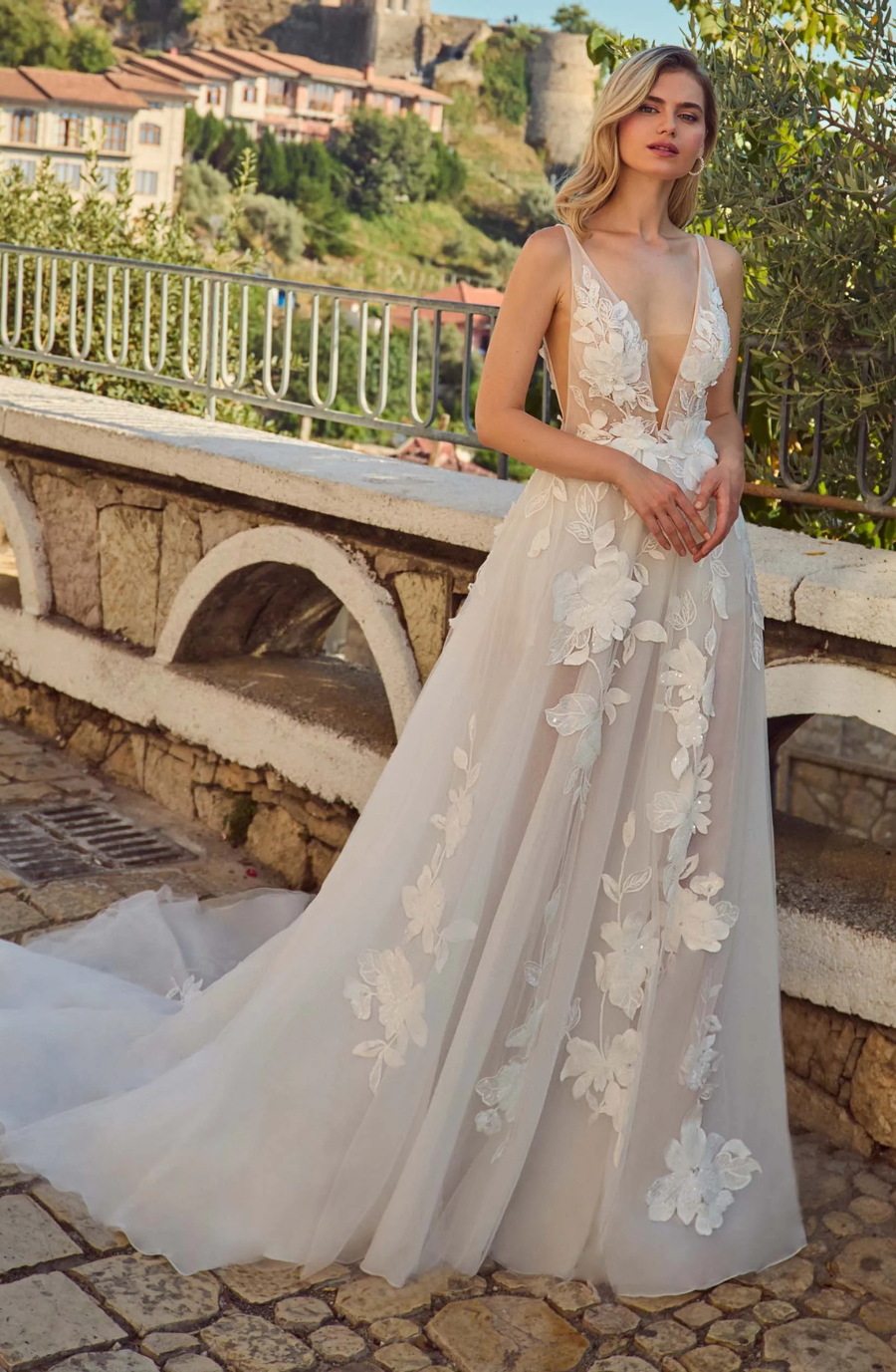 A woman in a white floral lace wedding dress stands outdoors by a stone railing, with a scenic background of trees and historic buildings.