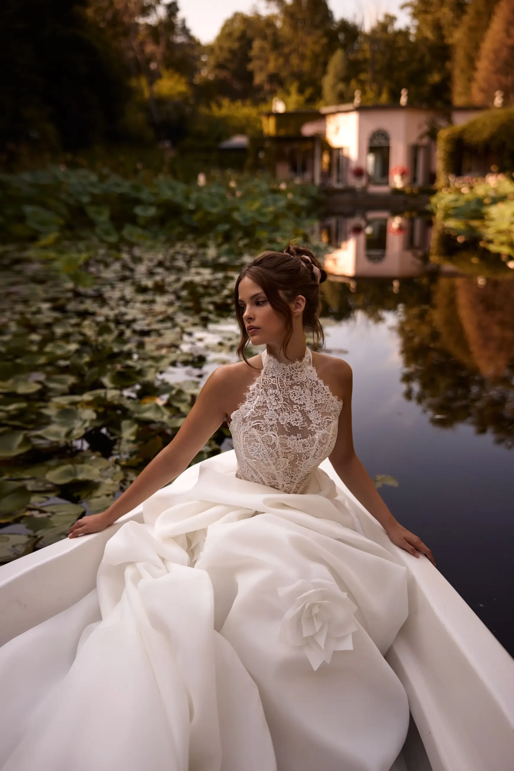 Model is wearing a halter-neck wedding gown with a fully embellished lace bodice, sheer boning, and a dramatic draped organza skirt that transforms into a high-low style.