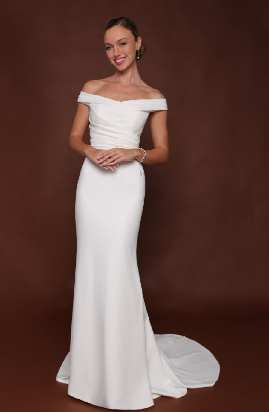 A woman in an off-the-shoulder, floor-length white gown stands against a dark brown background, smiling warmly. Her hair is elegantly styled back.