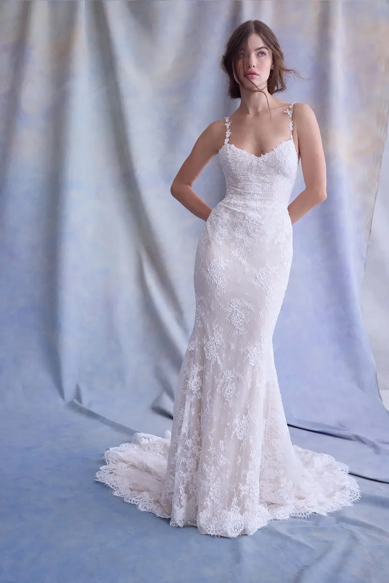 A woman in a fitted lace wedding gown stands against a soft blue and beige backdrop. Her expression is serene, and the dress has intricate floral patterns.