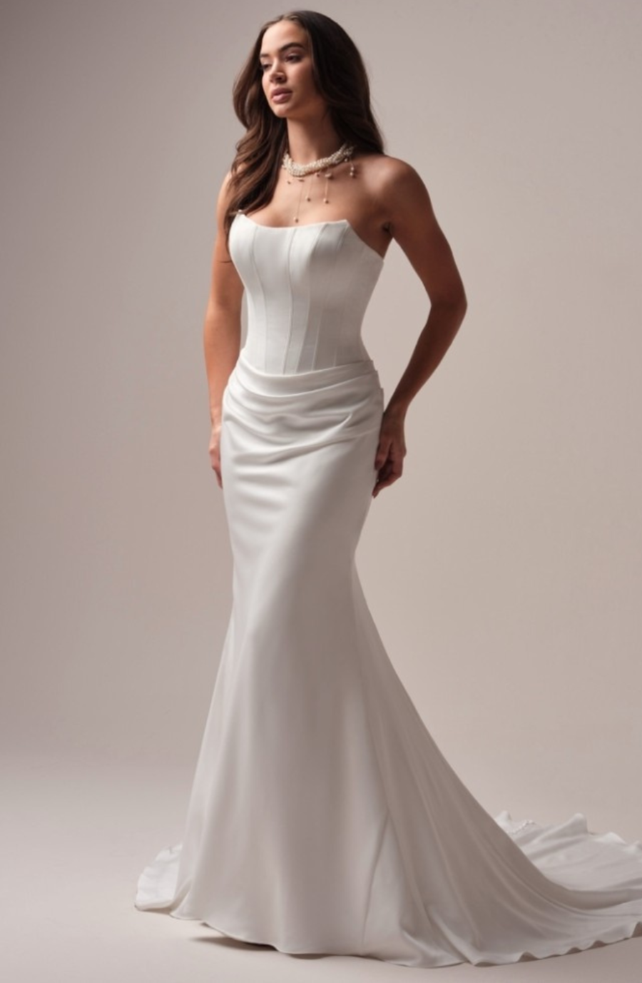 A woman in an elegant white strapless wedding gown with a fitted bodice, flowing skirt, and pearl necklace stands gracefully against a soft, neutral background.