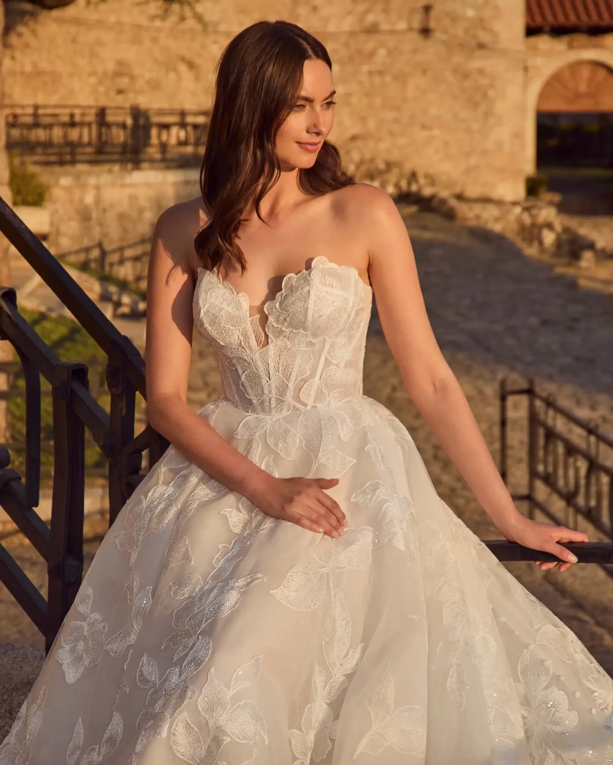 Model is wearing a strapless ball gown with a structured floral lace corset bodice and full tulle skirt.