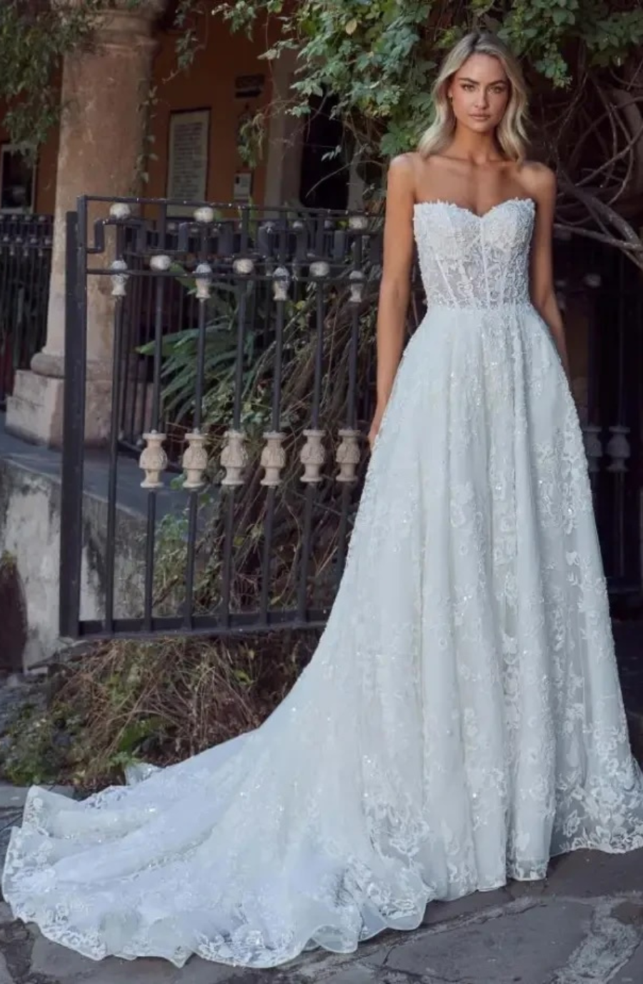 A bride in a strapless, lace wedding gown stands on a stone path. The dress features a long train. She is in front of a wrought iron gate with greenery around, exuding elegance and romance.