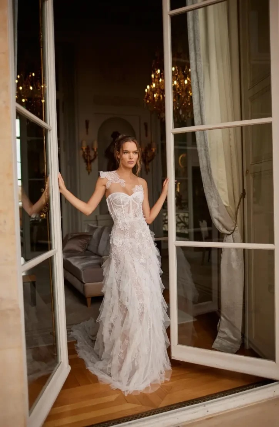 A woman in a delicate, lace wedding dress stands in an open French door. The room behind her is elegant, with soft lighting and a luxurious atmosphere.