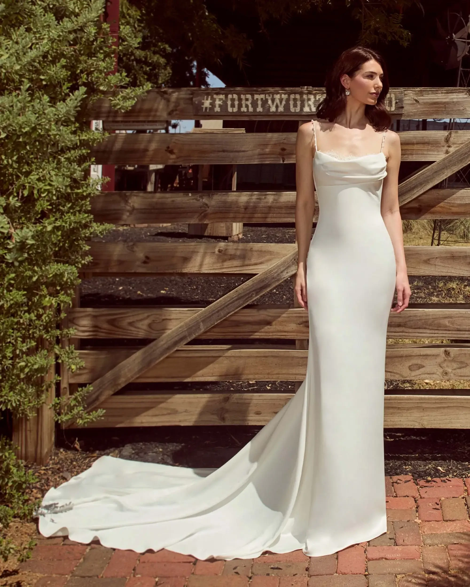 Model is wearing a sleeveless sheath wedding dress with spaghetti straps and a cowl neckline