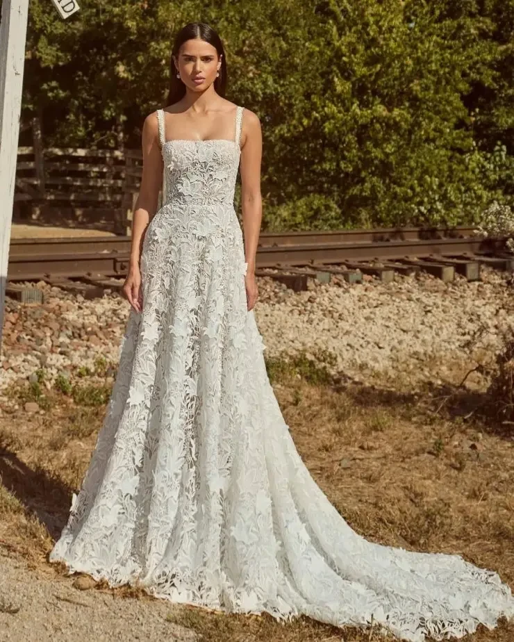 A model is standing outdoors in a flowing white lace dress with a fitted bodice and straps, surrounded by greenery and a gravel path.