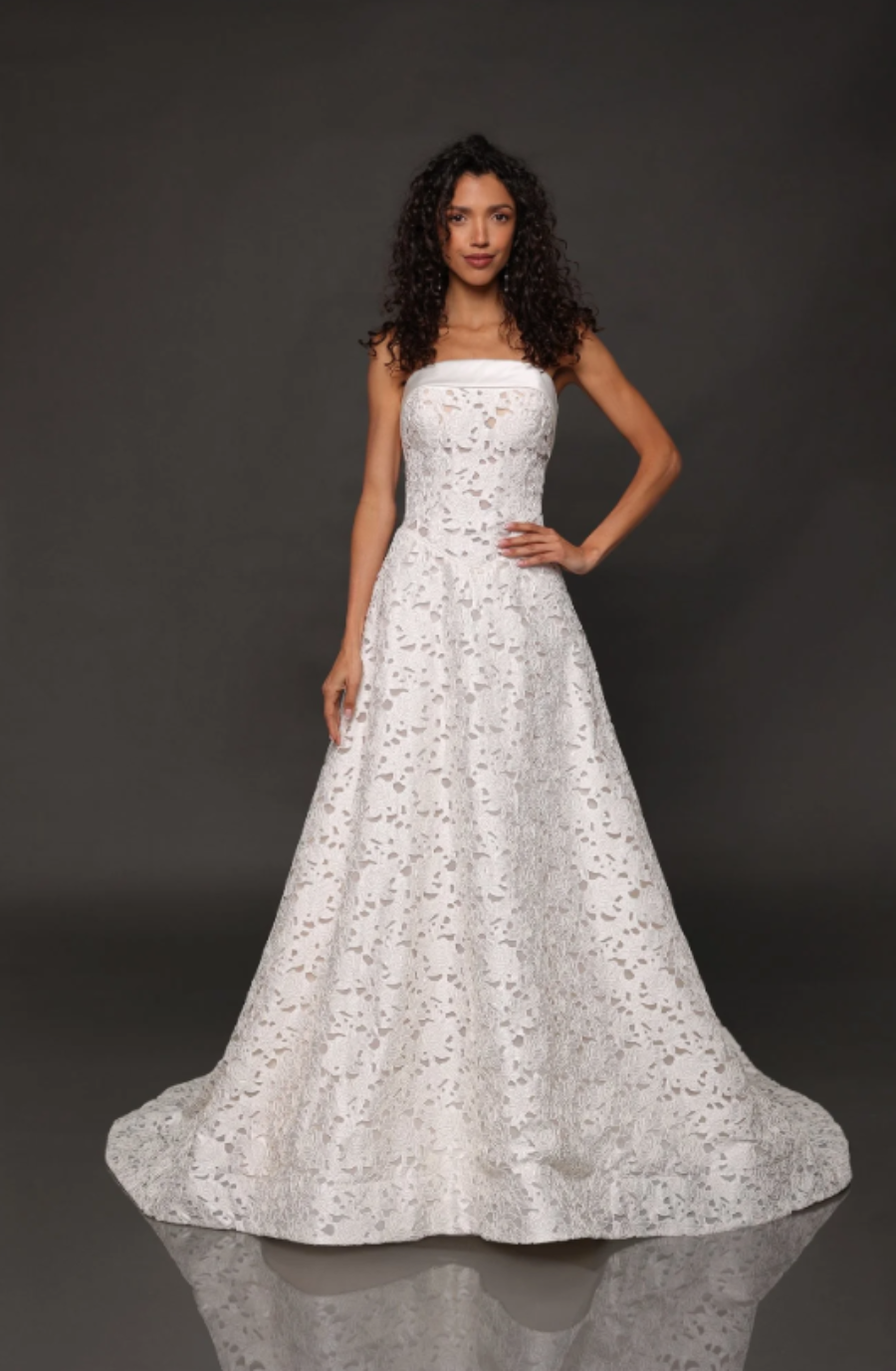 A woman poses confidently in a strapless, floor-length white lace gown against a dark backdrop. Her curly hair frames her face, projecting elegance.