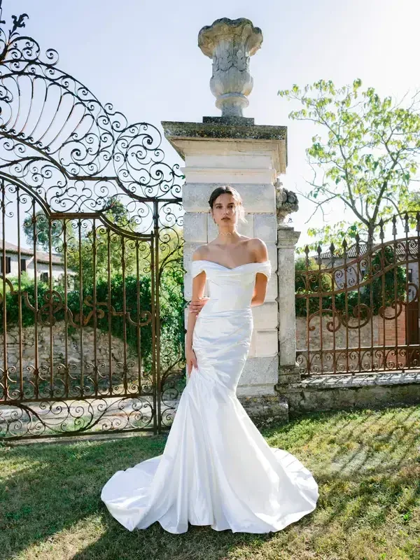 A model in a white off-the-shoulder wedding dress stands outside by an ornate gate, surrounded by greenery.
