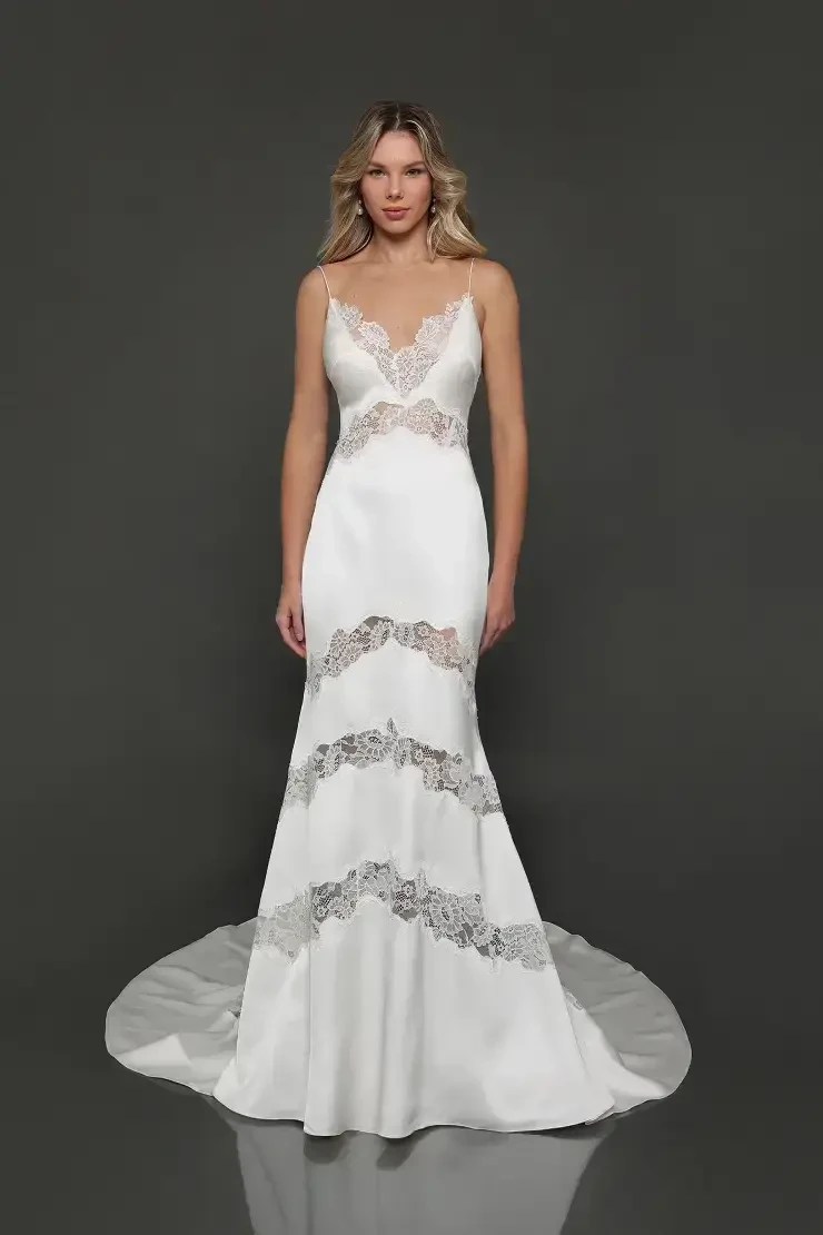 A model wearing a sleek, fitted wedding gown with a deep V-neckline and intricate lace detailing, standing against a dark background.