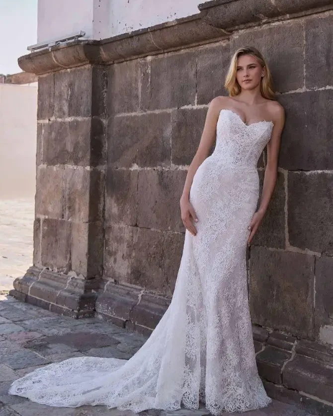A model wearing a strapless white lace wedding gown standing against a stone wall.