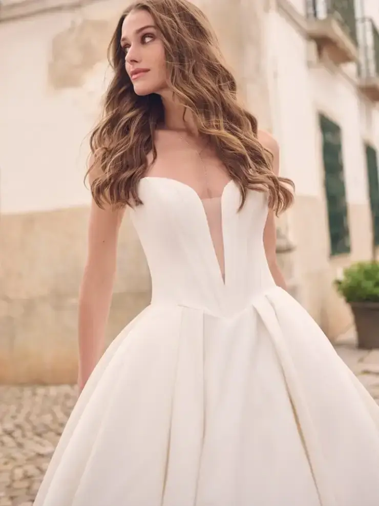 A woman in a strapless white wedding dress with a deep V-neckline stands on a cobblestone street, looking contemplative. Her long hair is styled in loose waves.