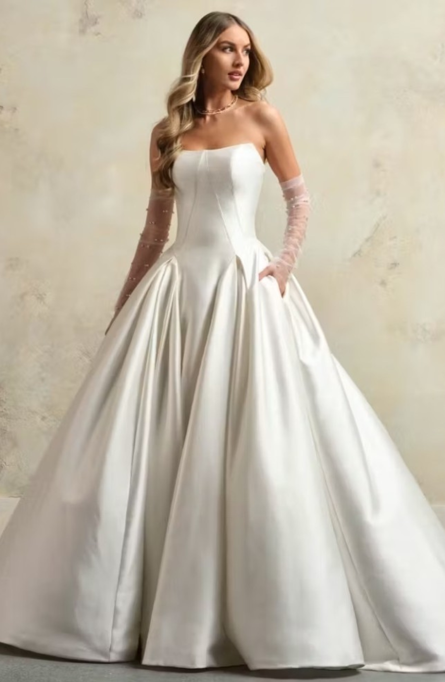 A woman in an elegant strapless white ball gown with sheer, embellished sleeves stands against a neutral background, exuding grace and sophistication.