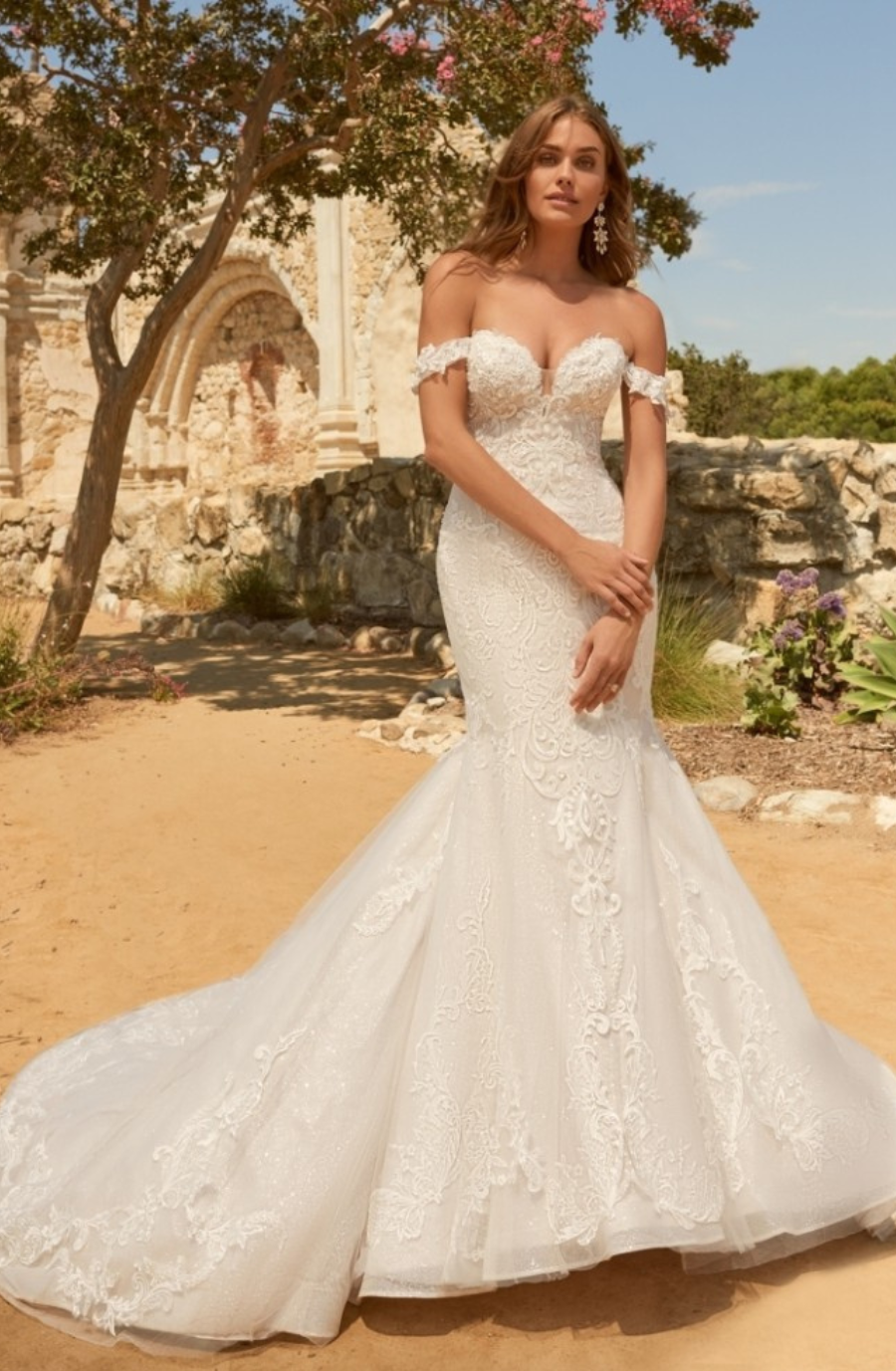 Bride in an elegant strapless lace wedding gown stands outside near stone ruins and a tree, with sunlight creating a romantic ambiance.