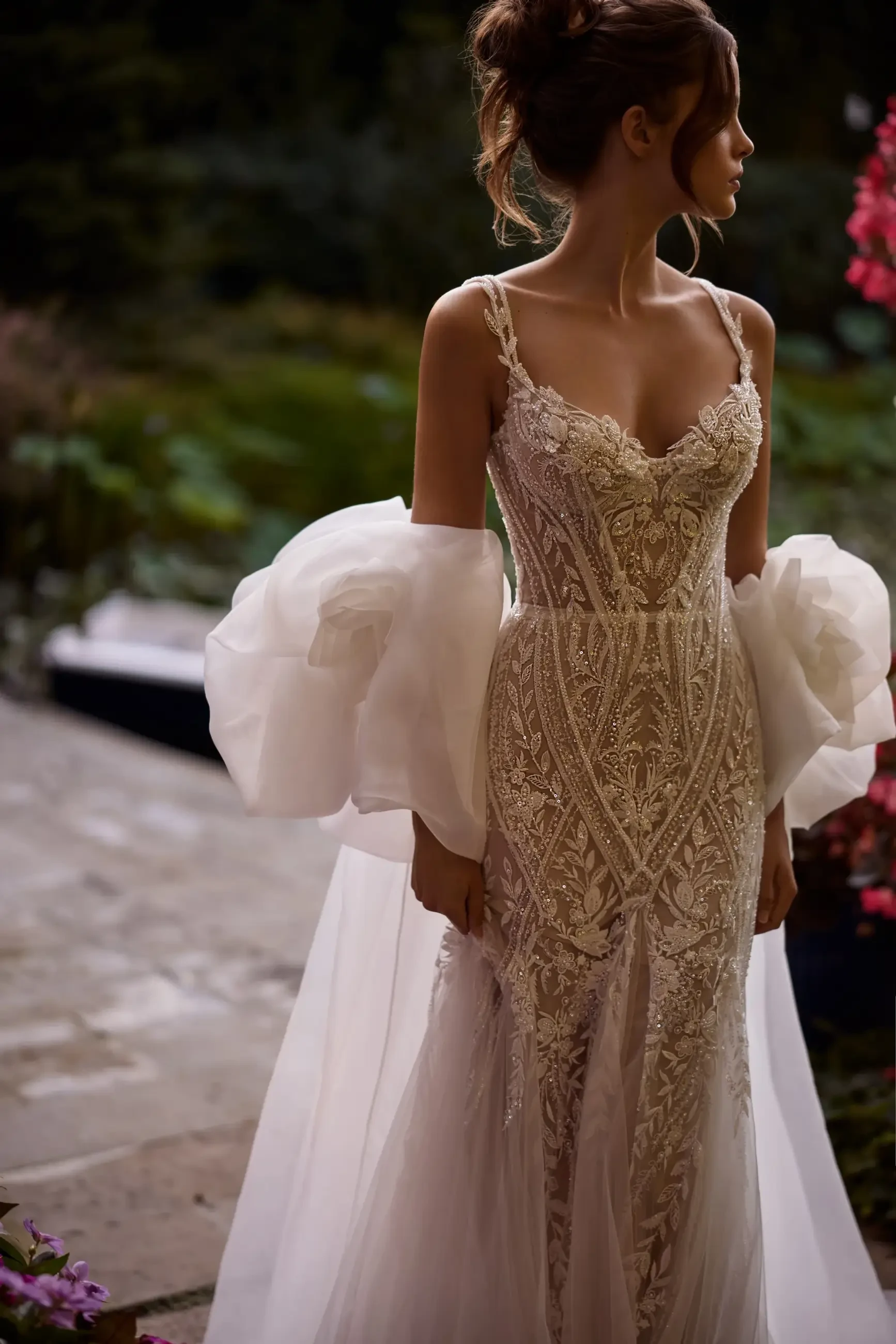A woman in an ornate, lace wedding gown with delicate straps stands outdoors. The dress is detailed with intricate embroidery. Her hair is styled up, and she exudes elegance.