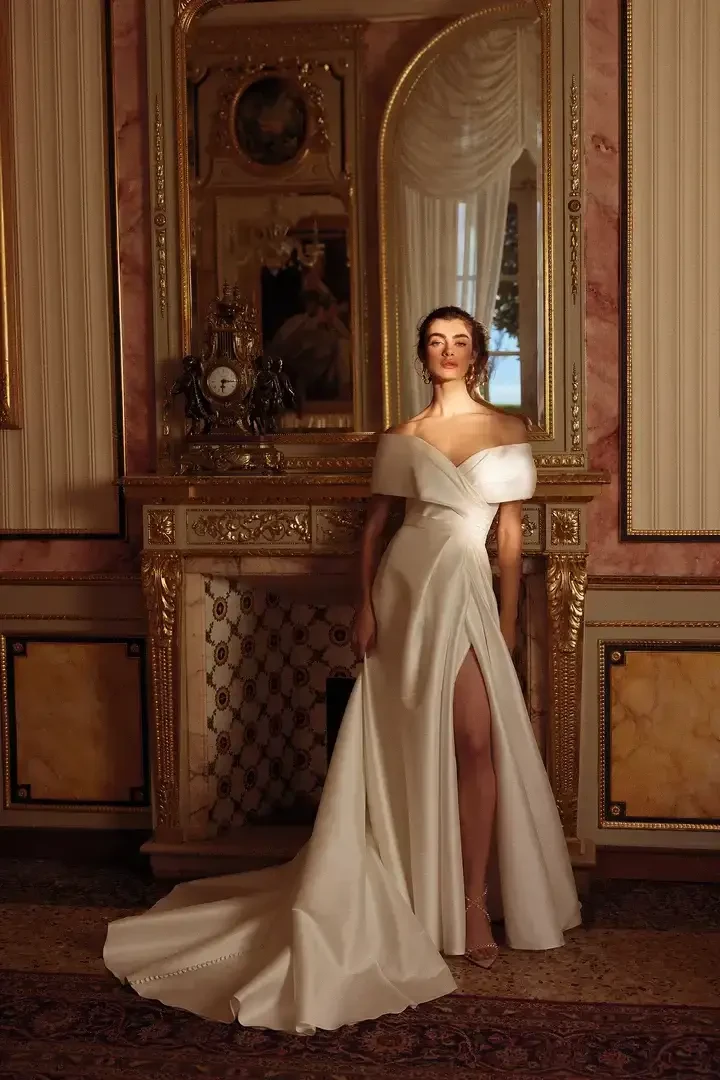 A woman in an elegant white gown stands in a lavishly decorated room with a large mirror and ornate features.