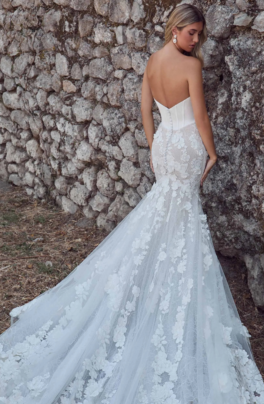 A woman in a strapless lace wedding gown with floral patterns stands beside a rustic stone wall. The dress has a long train, creating an elegant, romantic feel.