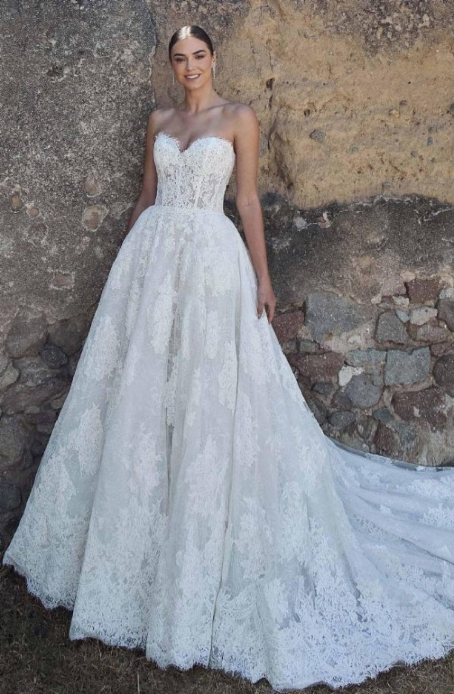 Elegant bride in strapless, lace wedding gown stands by a rustic stone wall. She smiles subtly, exuding a serene and joyful ambiance.