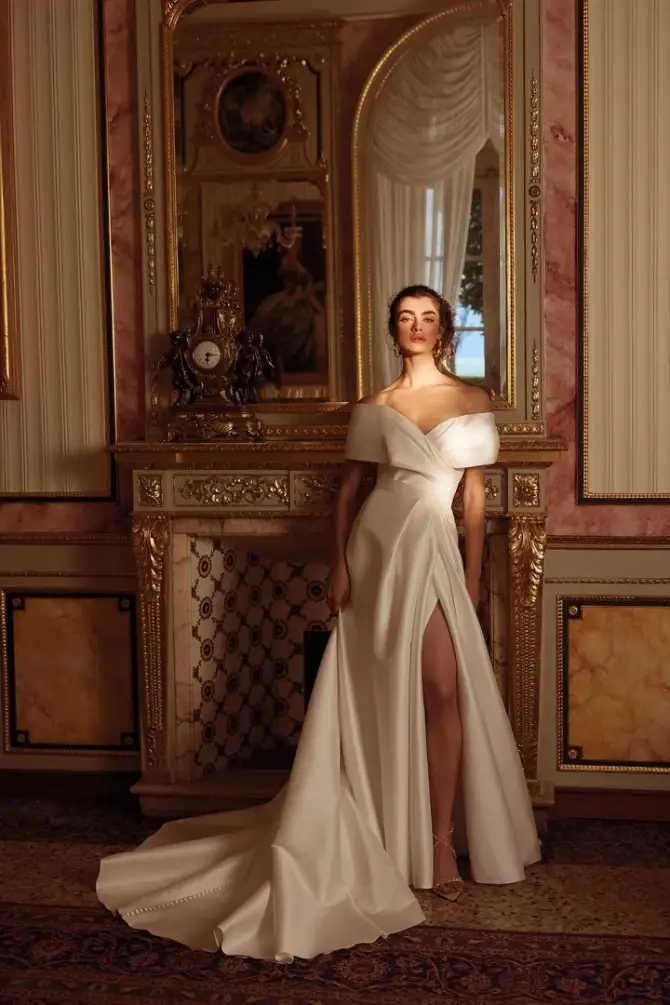 A woman wearing an elegant white gown stands in a lavish room with a large mirror and ornate decor.