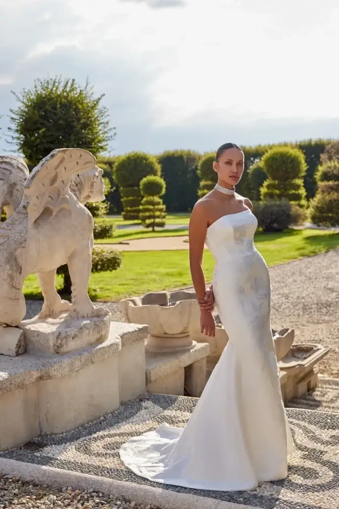 A woman in a strapless white wedding gown stands elegantly beside a stone sculpture in a beautifully landscaped garden.