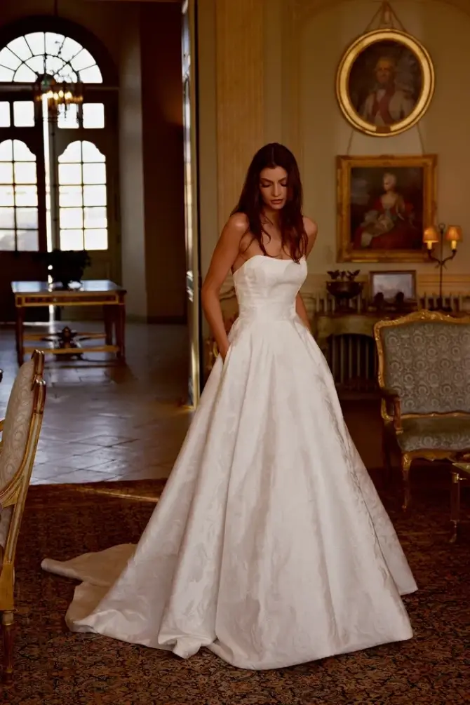 A woman in a strapless white wedding gown stands elegantly in a beautifully decorated room with vintage furniture and large windows.
