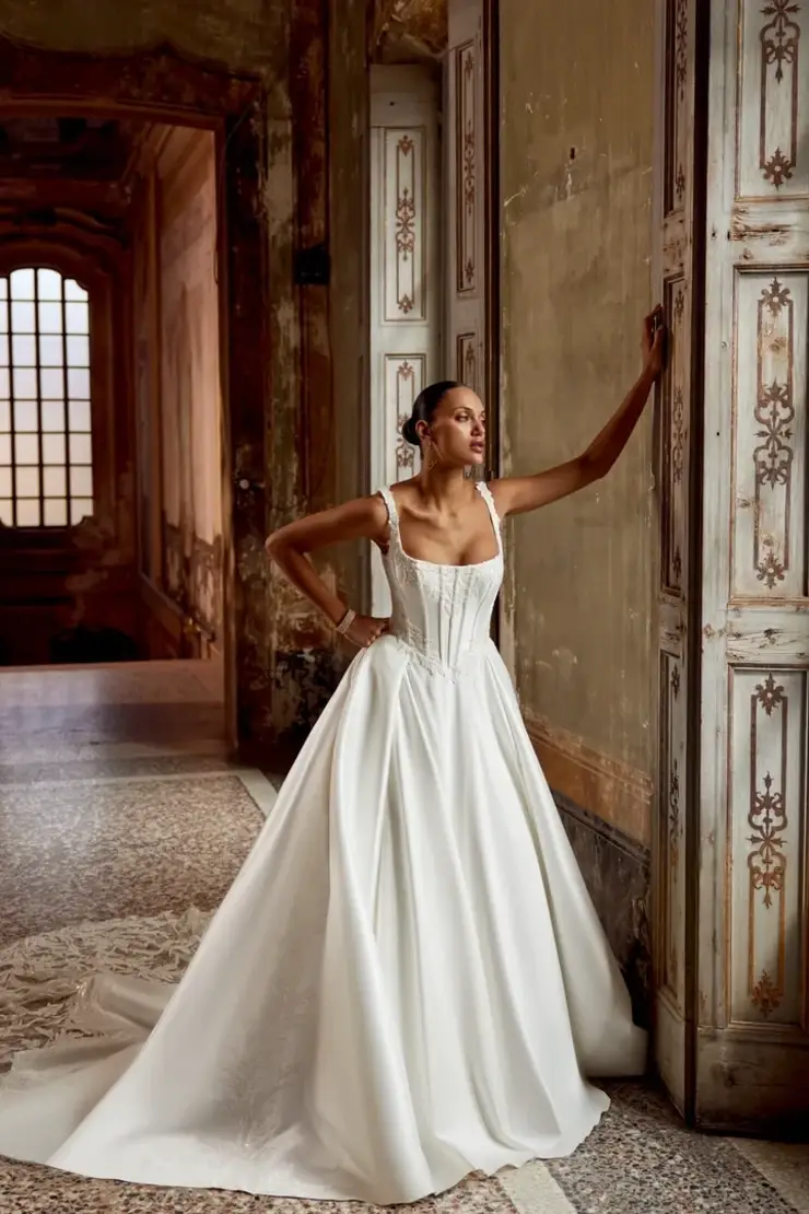 A woman wearing a white wedding dress poses elegantly against a wall in an ornate room.