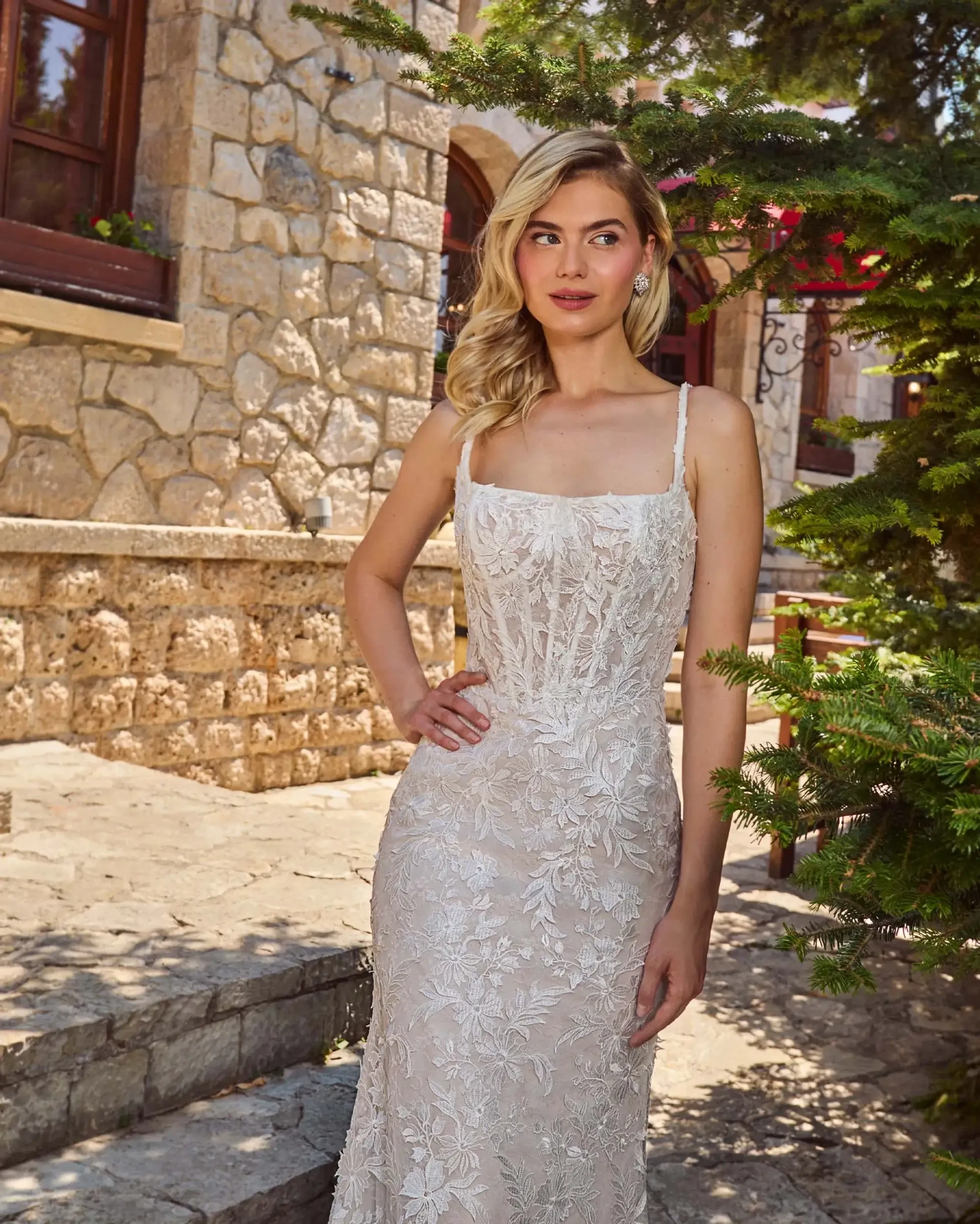 A woman in an elegant, floral lace gown poses outdoors near a stone building, exuding confidence. The setting is sunlit, creating a warm and serene atmosphere.