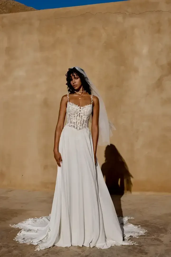 A model wearing a white wedding dress with a fitted bodice and a flowing skirt, standing against a textured beige wall, with a veil and soft curls.